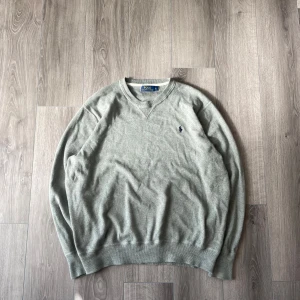 Grå tröja från Polo Ralph Lauren - Säljer en stilren grå tröja från Polo Ralph Lauren med v-ringning och broderad logga på bröstet. Perfekt för en avslappnad och snygg look. Tröjan har långa ärmar och är tillverkad i ett mjukt material.