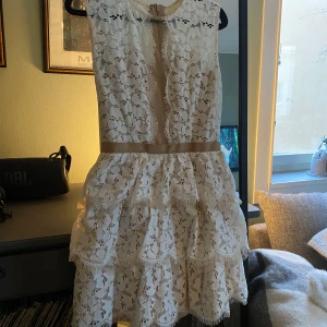 Vit spetsklänning med volanger - Charmig vit spetsklänning med volanger och beige detaljer. Klänningen är ärmlös och har en elegant design med ett bälte i midjan. Perfekt för sommarens festligheter. Köptes för 1000kr säljer lite billigare då jag har haft den väldigt länge men aldrig haft möjlighet att använda.