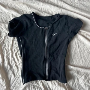 Svart kortärmad topp från Nike - Säljer en svart kortärmad topp från Nike med dragkedja framtill och vit piping längs kanterna. Perfekt för en sportig look eller avslappnad stil. Nike-loggan är diskret placerad på bröstet.