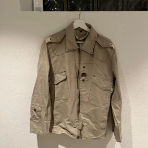 Beige jacka från HS - Säljer en beige jacka från HS med dragkedja och knappar. Jackan har långa ärmar och flera fickor framtill. Den har en stilren design med detaljerade sömmar på baksidan. Sitter ganska kort men bra