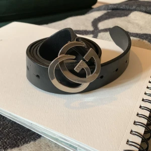 Svart skärp med silverdetaljer - Snyggt svart skärp  med en iögonfallande silverfärgad dubbel-G-spänne. Perfekt för att ge en lyxig touch till din outfit. Skärpet har flera hål för justerbar passform. Äkta läder. 