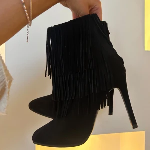 Svarta klackar med fransar - Snygga svarta boots med hög klack och fransdetaljer. Perfekta för att ge en trendig touch till din outfit. Tillverkade i mocka för en lyxig känsla. Väldigt bra skick men vid dragkedjan på ena skon är de lite trådar som hänger men de är bara att klippa av.