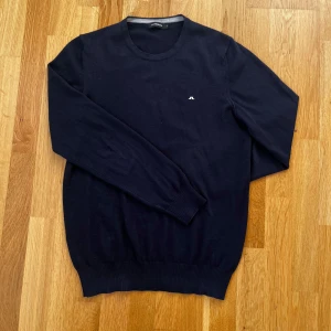 Mörkblå crewneck från J.Lindeberg - Säljer en stilren mörkblå tröja från J.Lindeberg med rund halsringning och diskret logga på bröstet. Perfekt för en klassisk look. Tröjan har långa ärmar och ribbade muddar vid ärmslut och nederkant. Det är ett litet litet hål i mitten samt en lagning på ena ärmen. Båda dessa syns ej tydligt!