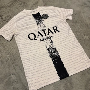 Vit fotbollströja med svart tryck - Säljer en vit fotbollströja från Nike med svart tryck och sponsorlogga. Tröjan har korta ärmar och ett unikt mönster i svart längs mitten. Perfekt för fotbollsfans!