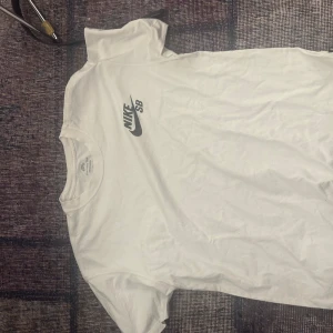 Vit t-shirt från Nike - Säljer en klassisk vit t-shirt från Nike med deras ikoniska logga 'Nike SB' tryckt på bröstet. Perfekt för en avslappnad stil. T-shirten är kortärmad och gjord i mjuk bomull för extra komfort.