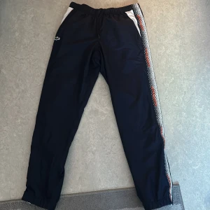 Mörkblå/svarta träningsbyxor från Lacoste Sport - Snygga svarta träningsbyxor från Lacoste Sport med vita och orange detaljer längs sidorna. Byxorna har en elastisk midja.