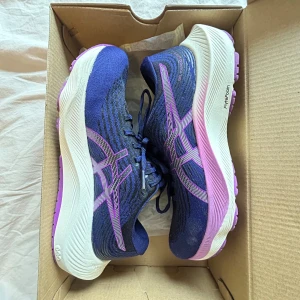Blå och lila löparskor från Asics - Snygga löparskor från Asics i en kombination av blått och lila med vita detaljer. Skorna har en bekväm passform och är perfekta för träning. De har snörning och en dämpande sula för extra komfort.
