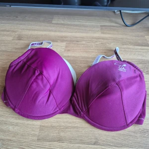 Helt ny bh med Snoopy-design 85C - Snygg vinröd bh med en söt Snoopy-design på kupan. De delvis glittriga axelbanden är justerbara och har Snoopy-text på sig. Uttagbara inlägg. Aldrig använd, prislapp kvar. Kommer från ett konkurslager. Se även mina andra auktioner för möjlighet till bundle med 20% rabatt.