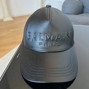 Svart keps från Balmain - Snygg svart keps från Balmain i skinn med broderad logga framtill. Kepsen har en justerbar rem baktill för perfekt passform. Perfekt för att ge din outfit en lyxig touch.
