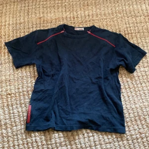 Svart t-shirt från Prada - Snygg svart t-shirt från Prada med röda dragkedjor på axlarna. Lite smått cropped också, priset kan diskuteras😊