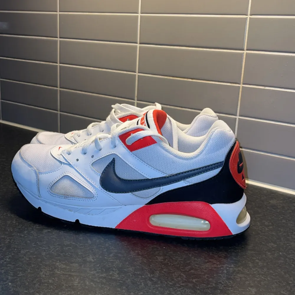 Säljer ett par Nike Air Max 90 sneakers i vitt med mörkblå och röda detaljer. Skorna har en synlig luftkudde i sulan och klassisk snörning. Perfekta för en sportig look! I princip nyskick.. Kengät.