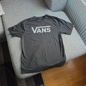 Svart t-shirt från Vans - Säljer en svart t-shirt från Vans med ett stort logotryck i grått mönstrat på bröstet. T-shirten har en klassisk passform och är kortärmad, perfekt för en avslappnad stil. Materialet känns mjukt och bekvämt.