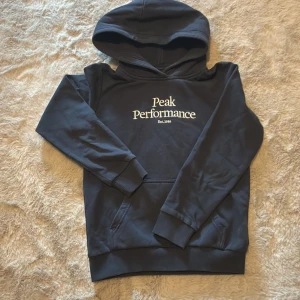 Mörkblå hoodie från Peak Performance - Säljer en mörkblå hoodie från Peak Performance med vit text på bröstet. Tröjan har en klassisk känguruficka och en bekväm huva. Perfekt för kyliga dagar!