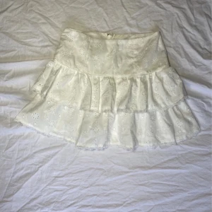 Vit kjol med spetsdetaljer - Charmig vit kjol med volanger och spetsdetaljer. Perfekt för en somrig look. Kjolen har en kort längd och en dragkedja baktill för enkel påklädning.