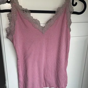 Rosa linnetopp med spetsdetaljer - Säljer en söt rosa ribbad linnetopp med grå spetsdetaljer längs kanterna. Toppen har justerbara axelband och en v-ringad design. Köpt för 50kr på new Yorker