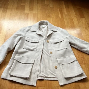 Vit jacka från H&M - Säljer en stilren vit jacka från H&M med knappar framtill och flera praktiska fickor. Jackan har en klassisk krage och är perfekt för vår och sommar. Den är tillverkad i ett mjukt material som ger en bekväm passform.
