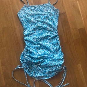 Blommig blå klänning stl XS - Söt blå klänning med blommigt mönster och smala axelband. Klänningen har en åtdragbar detalj på sidan som ger en snygg rynkad effekt. Perfekt för sommarens varma dagar!