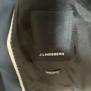 Midnattsblå kavaj från J.Lindeberg - Elegant midnattsblå kavaj från J.Lindeberg i regular fit. Kavajen har två knappar framtill och klassiska slag. Ärmarna är långärmade med en stilren randig insida. Perfekt för en stilren look.