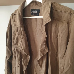 Grön trenchcoat från Bershka - Säljer en snygg grön trenchcoat från Bershka i 100% lyocell. Jackan har långa ärmar och knappar på ärmsluten som kan vikas upp för en ny look. Mjukt tyg och trendig design. Bältet har fått fötter så det följer inte med. Litet slitage på flärpen på baksidan men det syns inte 💛 
