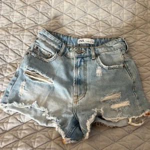 Blå  jeansshorts från Zara - Säljer ett par coola blå jeansshorts från Zara med slitna detaljer och fransar. Shortsen har en klassisk femficksdesign och knappgylf. Perfekta för en avslappnad sommarlook!