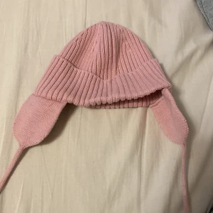 Rosa stickad mössa från Lindex - Söt rosa stickad mössa från Lindex med öronlappar och knytband. Perfekt för att hålla små huvuden varma under kyliga dagar. Ribbad design ger extra stil och komfort.