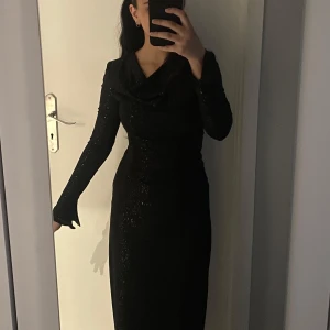Svart glittrig långklänning - Elegant svart långklänning med glittrande detaljer. Klänningen har långa ärmar och en draperad halsringning som ger en sofistikerad look. Perfekt för festliga tillfällen.