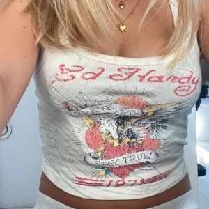 Ed Hardy x Urban Outfitters linne - Säljer ett vitt croppat linne från Ed Hardy x Urban Outfitters med ikoniskt tryck framtill. Linnet har ett stort hjärta, örn och texten 'Stay True' i rött och svart. Snyggt broderat Ed Hardy-tryck upptill och breda axelband. Perfekt för dig som gillar statement-plagg!
