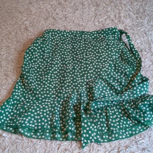 Grön blommig omlottkjol med volang - Supersöt grön kort omlottkjol med vita små blommor och volangkant. Kjolen har knytband i sidan och ett flowy fall som ger en riktigt somrig vibe. Perfekt för dig som gillar färg och mönster! 🌼