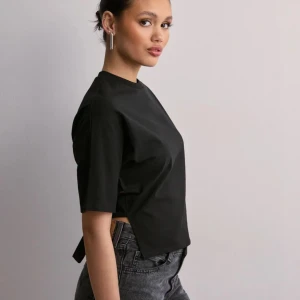 Svart croppad topp med knyt i ryggen - Trendig svart croppad topp med korta ärmar och snygg knytning i ryggen. Toppen har en rak passform framtill och öppen rygg baktill med breda band som knyts till en rosett. Perfekt att styla med höga jeans för en cool look.