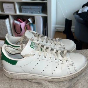 Adidas Stan Smith vita sneakers - Klassiska Adidas Stan Smith sneakers i vitt skinn med gröna detaljer på häl och plös. Skorna har rund tå, snörning och perforerade ränder på sidorna. Perfekt för en clean och tidlös stil.