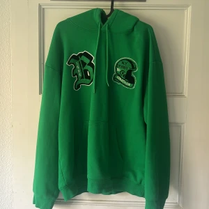 Grön hoodie med broderade patches - Snygg grön hoodie med broderade patches framtill och texten 'U.S. Football 76-79 League Champs 79' på ryggen. Klassisk känguruficka, snörning i huvan och oversized passform. Perfekt för dig som gillar sportig och avslappnad stil.