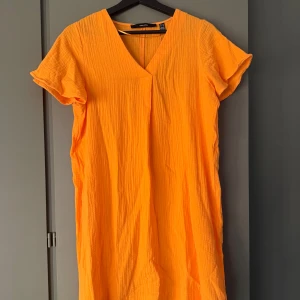 Orange kortklänning med v-ringning - Superfin orange kortklänning från Vero Moda  i ett luftigt bomullstyg. Klänningen har v-ringad hals, korta ärmar och en avslappnad passform. Perfekt strandklänking. Prislapp kvar 