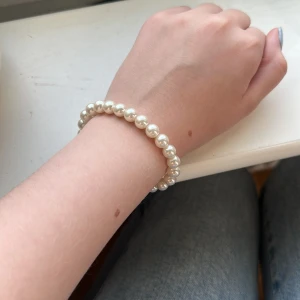 Pärlarmband med vita pärlor - Stilrent armband med glansiga vita pärlor på elastisk tråd. Perfekt accessoar för att ge din outfit en klassisk och elegant touch. Pärlorna är runda och jämnstora, vilket ger ett snyggt och tidlöst intryck.