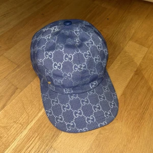 Blå keps från Gucci med monogram - Säljer en blå keps från Gucci med klassiskt GG-monogram över hela kepsen. Justerbar rem baktill i blått läder och insida i ljus bomull. Snygg och stilren accessoar som lyfter din outfit.
