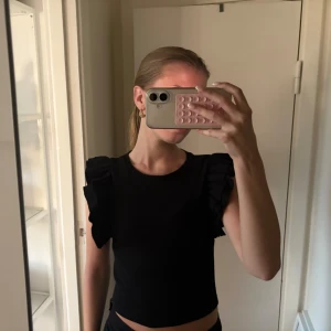 Svart  topp med volangärmar - Säljer en svart  topp med volangdetaljer på axlarna. Toppen har rund halsringning och är ärmlös med markerade volanger som ger en cool och trendig look. Storlek xs💕