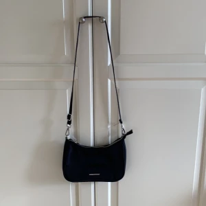 Svart axelväska från Bershka - Stilren svart axelväska från Bershka med justerbar rem och silvriga metalldetaljer. Väskan har en enkel, avlång form och stängs med dragkedja upptill. Tillverkad i syntetmaterial. I fint skick. 