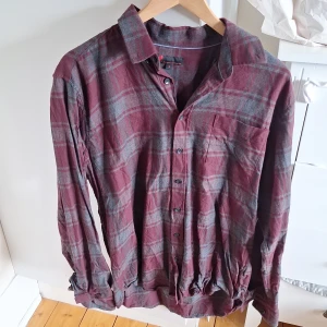 Rutig vinröd skjorta från Dressmann - Vinröd och grå rutig skjorta från Dressmann i storlek L. Skjortan har klassisk krage, knäppning framtill och en bröstficka. Långärmad modell i mjukt bomullstyg, perfekt för en avslappnad och snygg look.