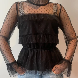 Svart meshblus med volanger och prickar - Supersnygg svart meshblus med volanger och prickigt mönster. Transparenta långärmar och hög hals ger en cool look. Blusen har resår i midjan och dekorativa spetsdetaljer vid axelbanden. Perfekt för dig som vill sticka ut lite extra.