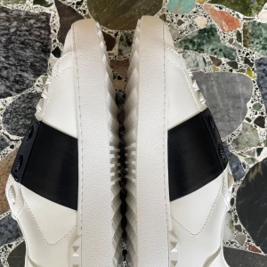 Valentino Garavani Open sneakers vit/svart 42 - Snygga Valentino Garavani Open sneakers i vitt skinn med bred svart läderdetalj över snörningen. Klassisk rund tå, platt sula med nitar och diskret logga på plösen och sulan. Perfekta för dig som gillar lyxiga och stilrena sneakers.