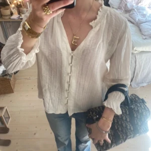 Vit blus med volangkant och knappar - Supersöt vit blus med volangkant vid hals och ärmslut. Djup v-ringning och klädda knappar framtill ger en romantisk vibe. Långärmad och i lätt, luftig bomull som passar perfekt till jeans eller kjol. !Första bilderna är tagna från en annan, sista är min!