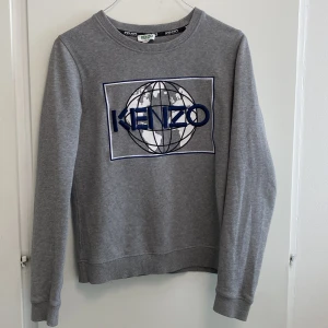 Grå Kenzo sweatshirt med broderi XS - Snygg grå sweatshirt från Kenzo i bomull med broderad världskarta och blått Kenzo-tryck på bröstet. Tröjan har rund hals, ribbade muddar och lång ärm. Perfekt för dig som gillar streetstyle och vill ha något bekvämt men ändå trendigt. ( vi kan absolut diskutera priser och jag kan såklart skicka fler bilder om det skulle behövas)🫶