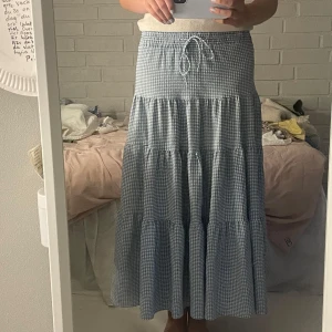 Blåvit rutig långkjol med snörning - Supersöt långkjol i blåvitt rutigt mönster med volanger och resår i midjan. Kjolen har snörning i midjan för extra passform och är gjord i ett lätt och luftigt bomullstyg. Perfekt för en chill sommardag eller picknick i parken.