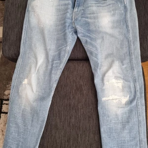 Replay anbass ljusa jeans med slitningar - Säljer ett par ljusblå jeans från Replay med snygga slitningar på benen och klassiska bakfickor med broderad detalj. Jeansen har normal passform och är tillverkade i bomull. Perfekta för en avslappnad och trendig look.