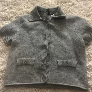 Grå stickad kofta från H&M, kortärmad - Kortärmad grå stickad kofta från H&M med ribbad krage, knappar framtill och två fickor med knappdetalj. Perfekt för lager-på-lager och chill stil. Mjuk och skön känsla, passar till många outfits. Fickorna går ej att öppna. Använd ungefär 2-3 gånger, säljer för ingen användning!💕