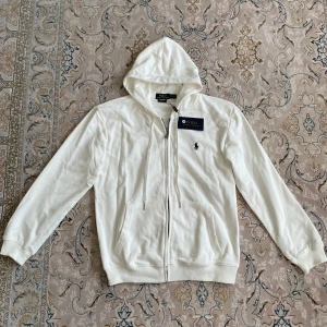 Vit hoodie från Polo Ralph Lauren M - Snygg vit hoodie från Polo Ralph Lauren i bomull med dragkedja och huva. Klassisk broderad logga på bröstet, två fickor framtill och snörning i huvan. Perfekt för en clean och stilren look. Custom fit-modell som sitter riktigt bra.