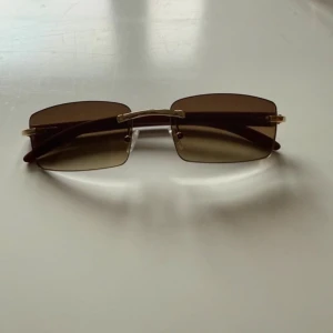 Bruna rektangulära solglasögon med guld - Snygga solglasögon med rektangulära, bruna tonade glas och guldfärgade detaljer. Skalmarna är bruna och har en stilren design utan båge runt glasen. Perfekt accessoar för en cool look i solen.