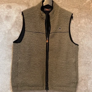 Olivgrön fleeceväst från Morris - Olivgrön fleeceväst från Morris med svart kantband och hel dragkedja framtill. Västen har hög krage, två fickor och en broderad logga på bröstet. Materialet är mjukt och fluffigt, perfekt för lager-på-lager under kyliga dagar.