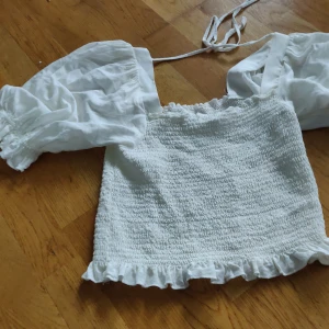 Vit blus med puffärmar och volang - Supersöt vit blus med smockad kropp, puffiga långärmar och volangkant nertill. Toppen har knytband upptill och är gjord i ett lätt bomullstyg. Perfekt för en romantisk look med trendiga detaljer.