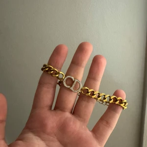 Guldigt CD-armband - armband i guldtonad metall med grova länkar och ett glittrande CD-emblem i mitten, täckt av små gnistrande stenar. Perfekt statement piece för dig som gillar lyxig och trendig stil. HELT OANVÄNT!