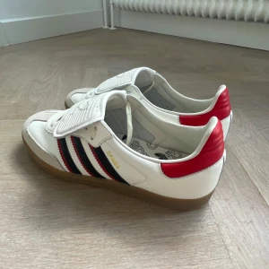Adidas Samba OG LT vit/röd sneakers - Adidas Samba OG sneakers i vitt skinn med röda detaljer på hälen och klassiska svarta och röda ränder på sidorna. Skorna har gummisula, snörning och en stor vit plös med Adidas-logga. Tidlös och sportig modell med retrovibbar. Tveka inte på att höra av dig!!!